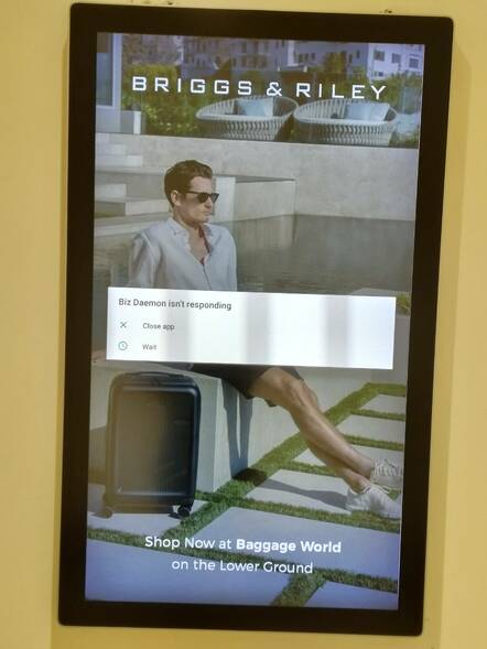 Electronic sign with pop-up text "Biz Daemon is not responding" over an image of a man in shorts sitting on a wall with a wheeled suitcase